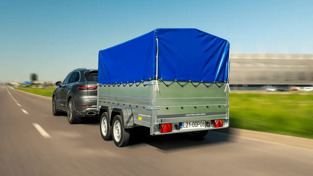 Szary SUV ciągnie przyczepkę z niebieską plandeką. W tle rozmyty krajobraz i budynek.