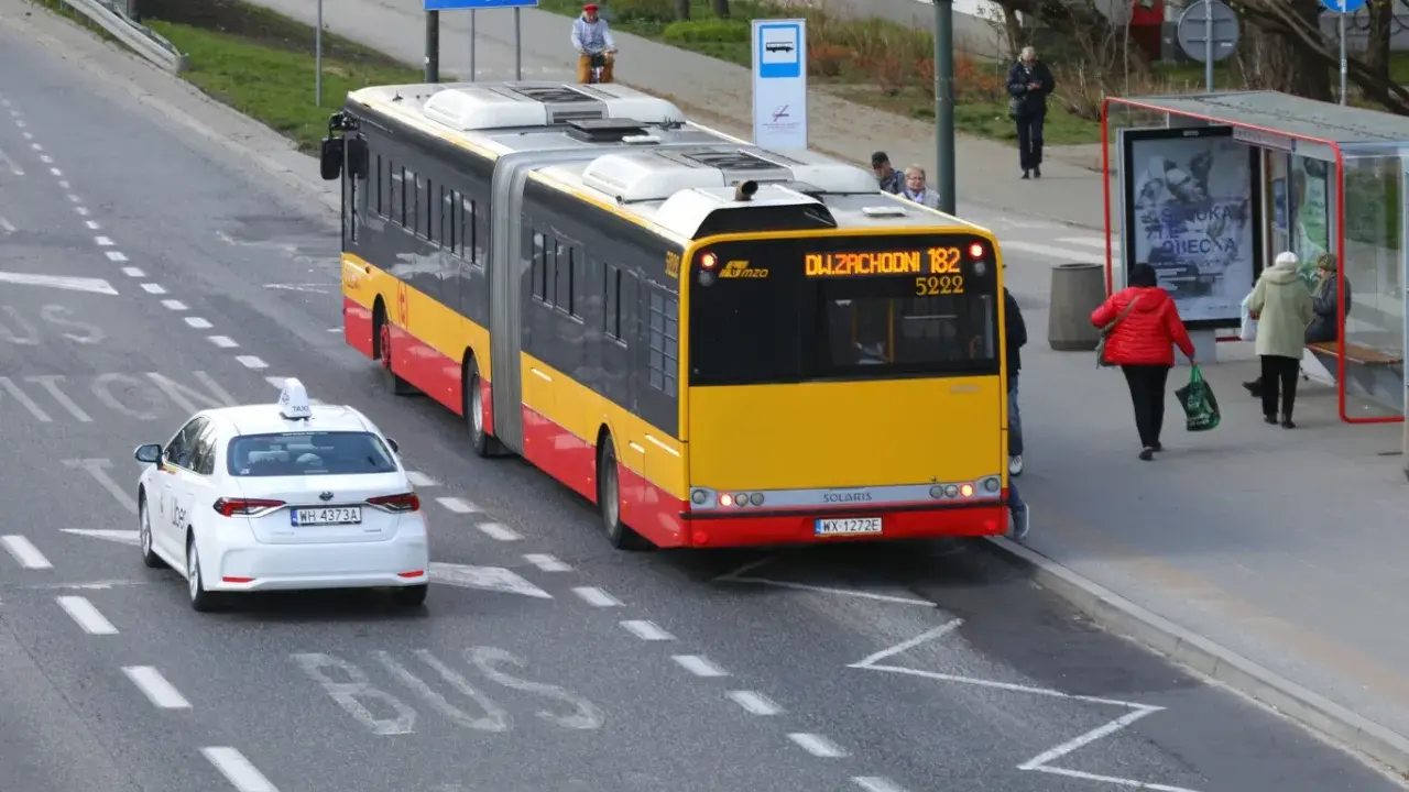 Żółto-czerwony autobus przegubowy linii 182 zbliża się do przystanku. Czy autobus ma pierwszeństwo przed białym autem taxi?