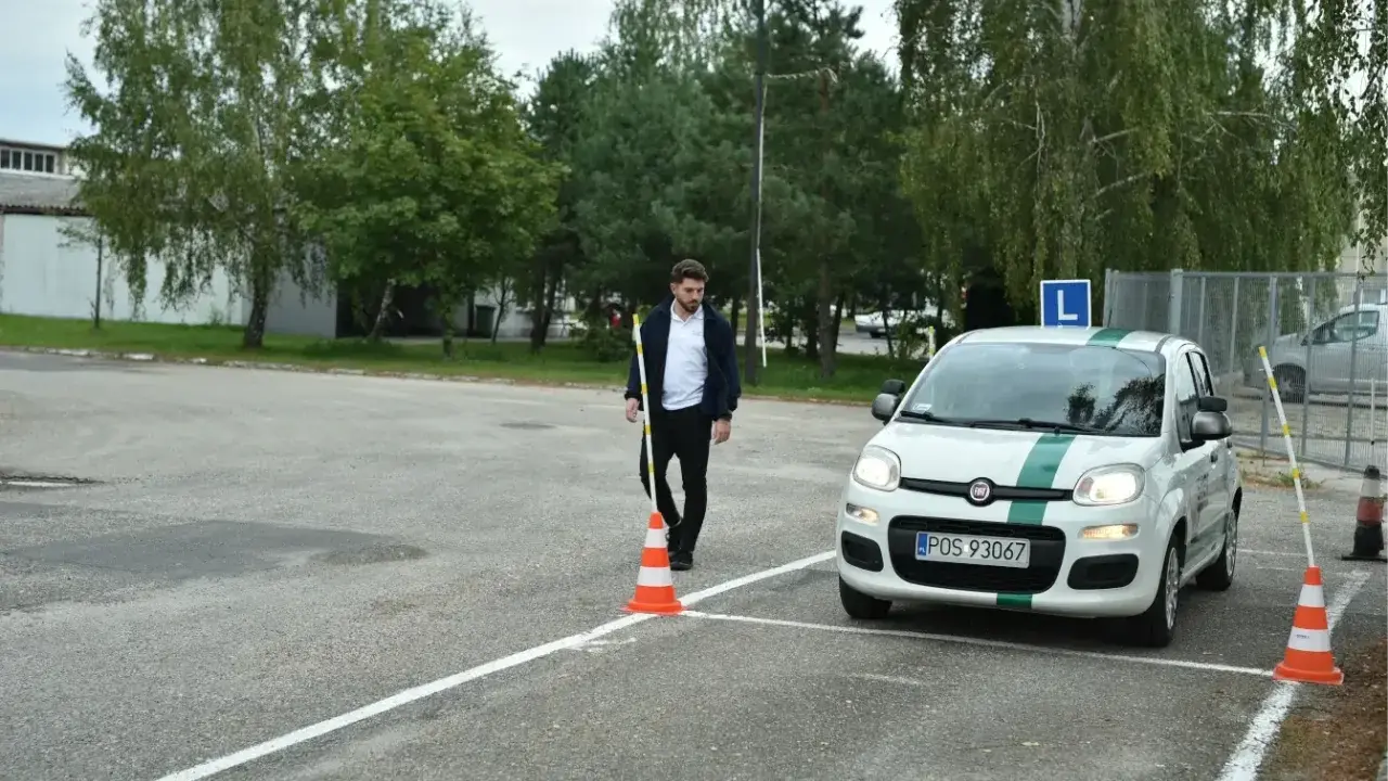 Młody mężczyzna na placu manewrowym, obok białego Fiata z zielonym pasem i literą L. Zapalone światła w samochodzie egzamin.