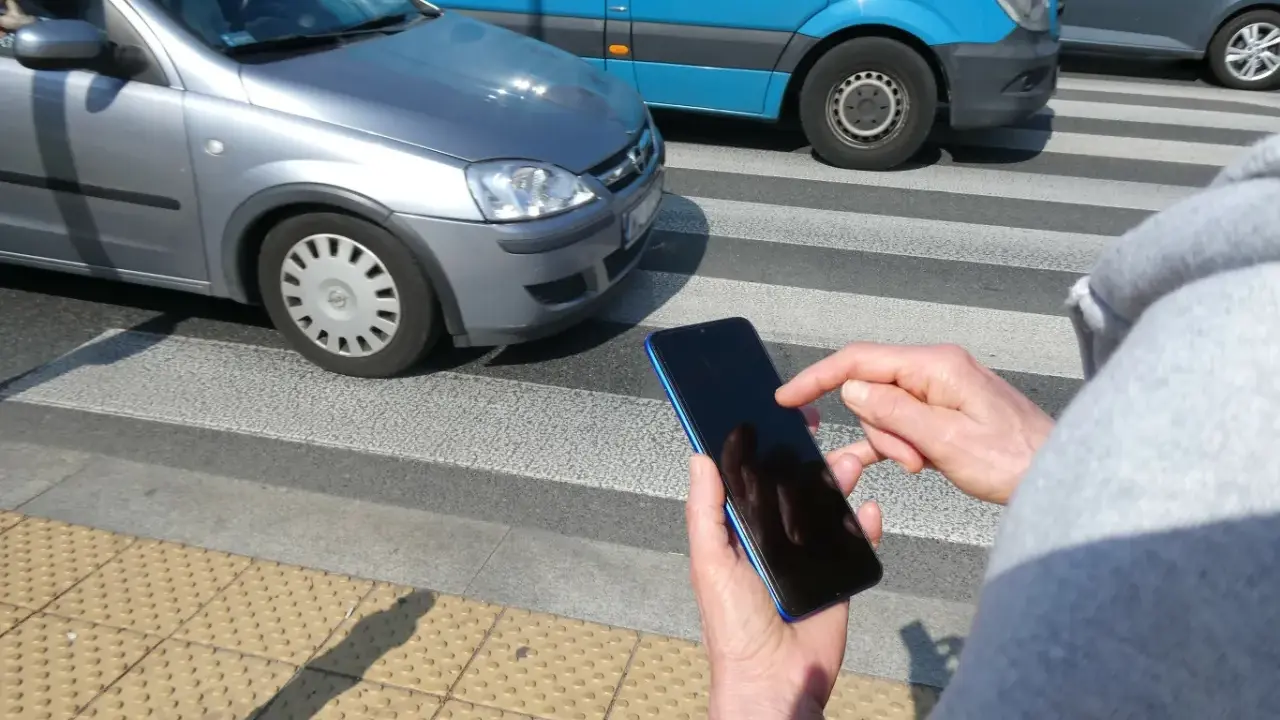 Osoba z telefonem na przejściu dla pieszych, kiedy należy zachować szczególną ostrożność. Samochody i niebieski van na drodze.