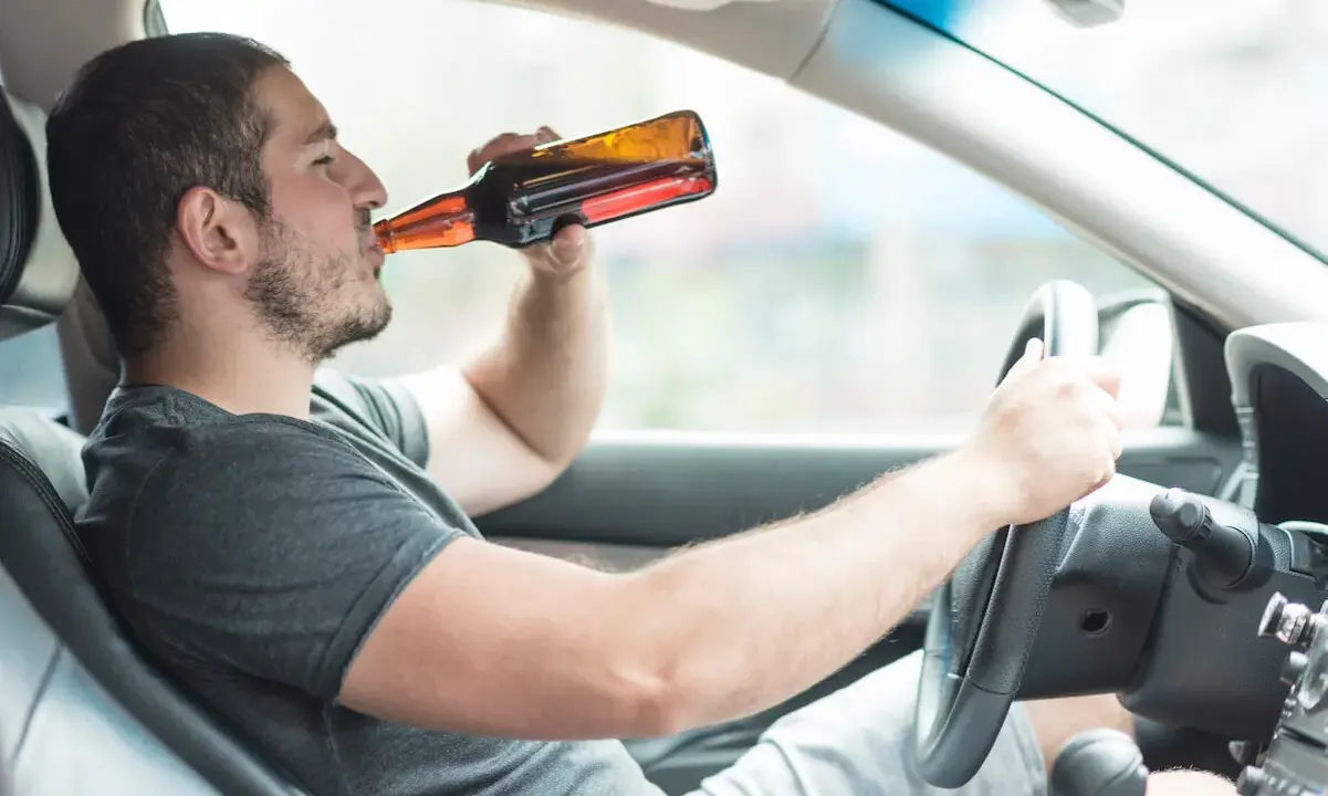 Mężczyzna pije piwo za kierownicą. Powyżej 0,5 promila jaka kara grozi za jazdę pod wpływem alkoholu.