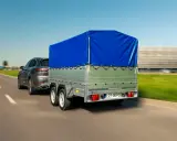 Szary SUV ciągnie przyczepkę z niebieską plandeką. W tle rozmyty krajobraz i budynek.