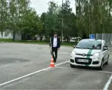 Młody mężczyzna na placu manewrowym, obok białego Fiata z zielonym pasem i literą L. Zapalone światła w samochodzie egzamin.