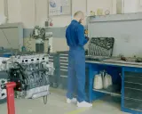 Mechanik w niebieskim kombinezonie przy silniku samochodu. Obok skrzynka z narzędziami i płyny. Lepiej zrobić przegląd, by uniknąć mandatu za brak przeglądu.