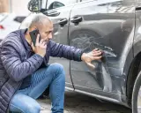 Mężczyzna sprawdza uszkodzenia samochodu po kolizji bez policji i oświadczenia, dzwoniąc z telefonu.