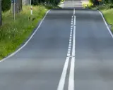 Droga z podwójną białą linią, która jest linia jednostronnie przekraczalna. Po bokach zieleń i drzewa.