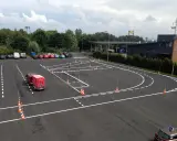 Plac manewrowy z wyznaczonymi trasami i pachołkami. Widać samochody, przyczepę kempingową i budynki w tle.