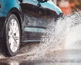 Koło czarnego samochodu wpada w kałużę, tworząc fontannę wody. To zjawisko to aquaplaning, czyli utrata przyczepności na mokrej nawierzchni.