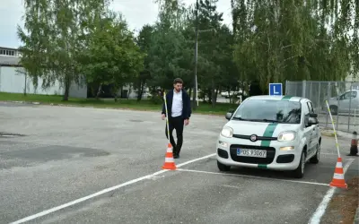 Młody mężczyzna na placu manewrowym, obok białego Fiata z zielonym pasem i literą L. Zapalone światła w samochodzie egzamin.