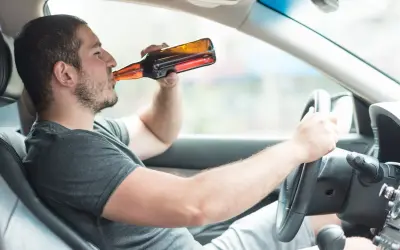 Mężczyzna pije piwo za kierownicą. Powyżej 0,5 promila jaka kara grozi za jazdę pod wpływem alkoholu.
