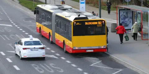 Żółto-czerwony autobus przegubowy linii 182 zbliża się do przystanku. Czy autobus ma pierwszeństwo przed białym autem taxi?