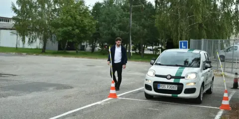 Młody mężczyzna na placu manewrowym, obok białego Fiata z zielonym pasem i literą L. Zapalone światła w samochodzie egzamin.