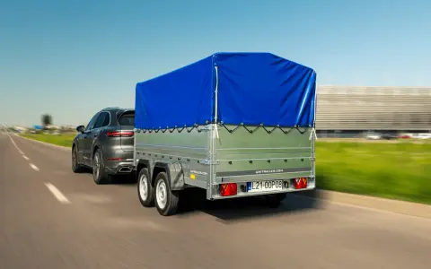 Szary SUV ciągnie przyczepkę z niebieską plandeką. W tle rozmyty krajobraz i budynek.