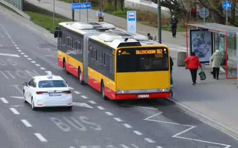 Żółto-czerwony autobus przegubowy linii 182 zbliża się do przystanku. Czy autobus ma pierwszeństwo przed białym autem taxi?