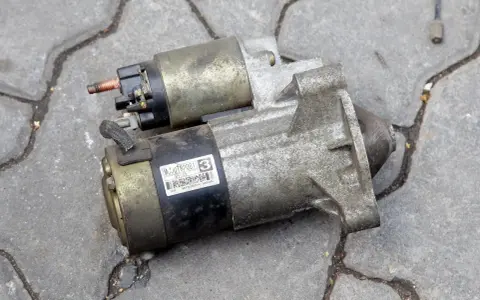 Dwa używane rozruszniki samochodowe, gotowe do wymiany. Jeden z nich ma naklejkę z numerem MC0JT82081.