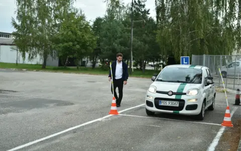 Młody mężczyzna na placu manewrowym, obok białego Fiata z zielonym pasem i literą L. Zapalone światła w samochodzie egzamin.