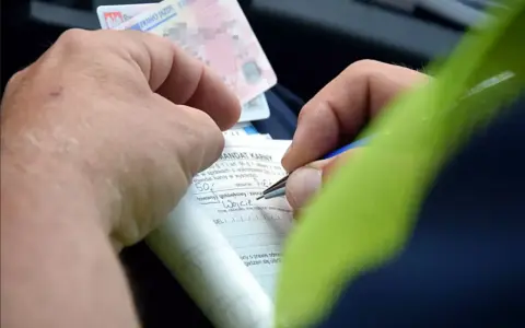Ręce wypełniają formularz mandatu karnego, próbując dokonać przepisanie mandatu na inną osobę. Widoczne prawo jazdy.
