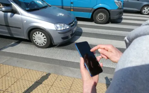 Osoba z telefonem na przejściu dla pieszych, kiedy należy zachować szczególną ostrożność. Samochody i niebieski van na drodze.