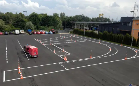 Plac manewrowy z wyznaczonymi trasami i pachołkami. Widać samochody, przyczepę kempingową i budynki w tle.