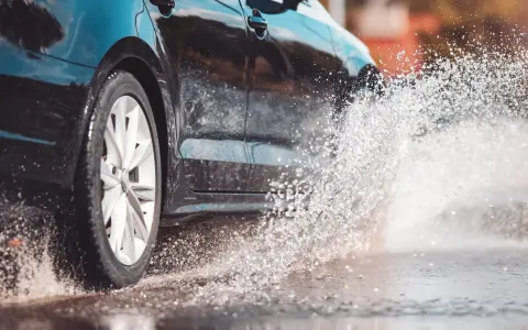 Koło czarnego samochodu wpada w kałużę, tworząc fontannę wody. To zjawisko to aquaplaning, czyli utrata przyczepności na mokrej nawierzchni.