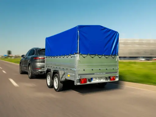 Szary SUV ciągnie przyczepkę z niebieską plandeką. W tle rozmyty krajobraz i budynek.