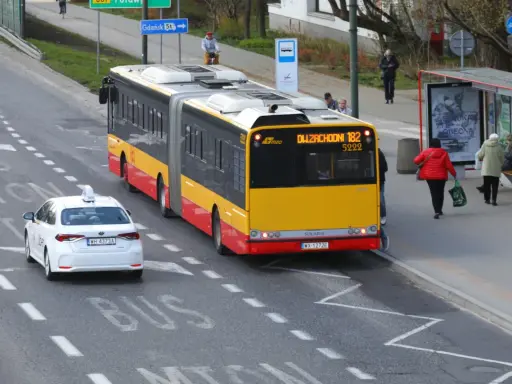 Żółto-czerwony autobus przegubowy linii 182 zbliża się do przystanku. Czy autobus ma pierwszeństwo przed białym autem taxi?