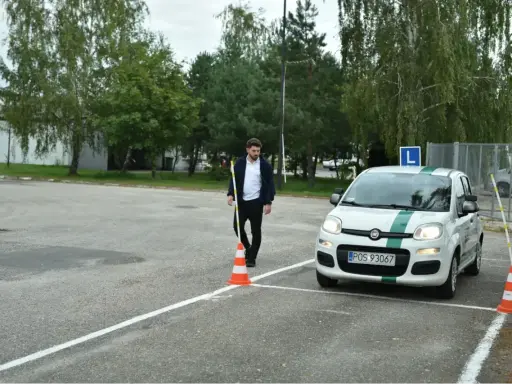 Młody mężczyzna na placu manewrowym, obok białego Fiata z zielonym pasem i literą L. Zapalone światła w samochodzie egzamin.