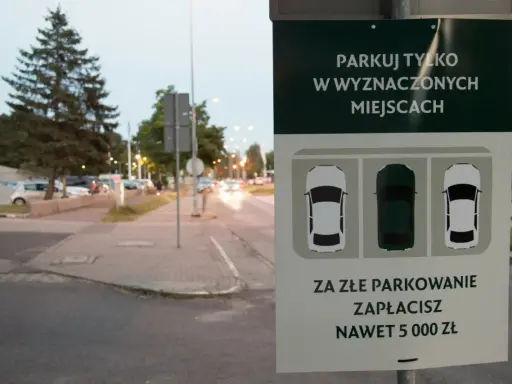 Znak informuje: "Parkuj tylko w wyznaczonych miejscach. Za złe parkowanie zapłacisz nawet 5000 zł". Straż miejska pilnuje porządku.