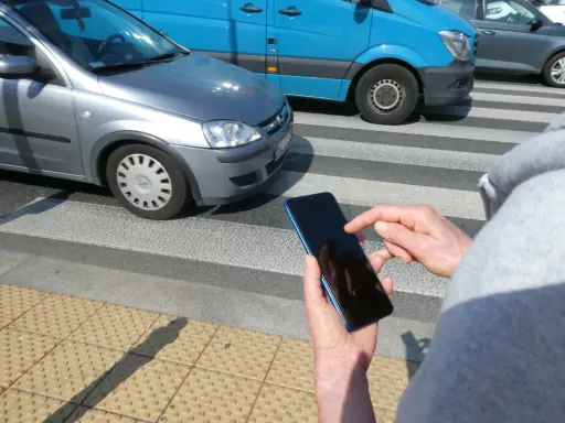 Osoba z telefonem na przejściu dla pieszych, kiedy należy zachować szczególną ostrożność. Samochody i niebieski van na drodze.