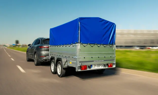 Szary SUV ciągnie przyczepkę z niebieską plandeką. W tle rozmyty krajobraz i budynek.
