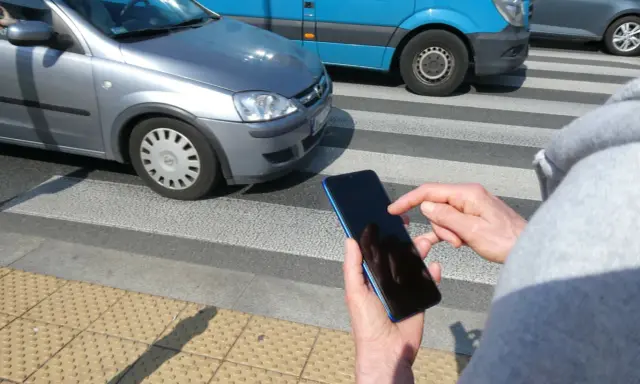 Osoba z telefonem na przejściu dla pieszych, kiedy należy zachować szczególną ostrożność. Samochody i niebieski van na drodze.