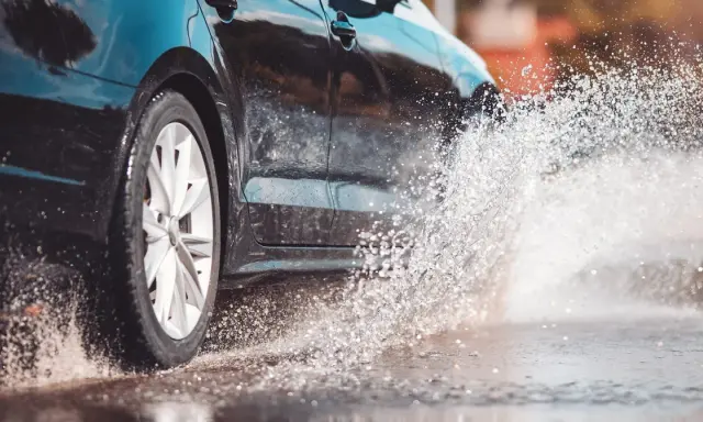 Koło czarnego samochodu wpada w kałużę, tworząc fontannę wody. To zjawisko to aquaplaning, czyli utrata przyczepności na mokrej nawierzchni.