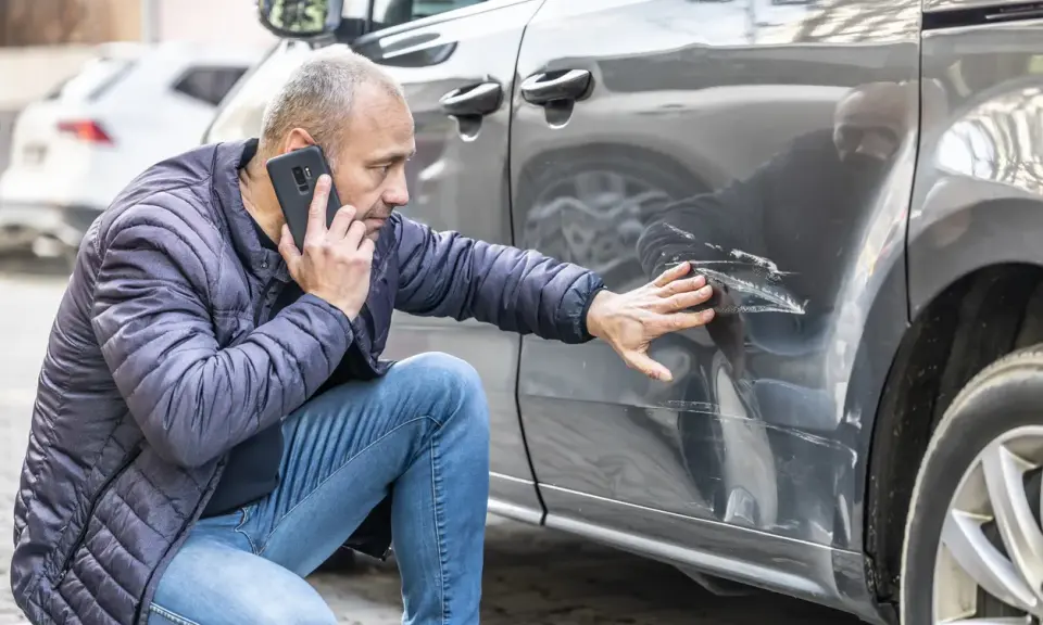 Mężczyzna sprawdza uszkodzenia samochodu po kolizji bez policji i oświadczenia, dzwoniąc z telefonu.