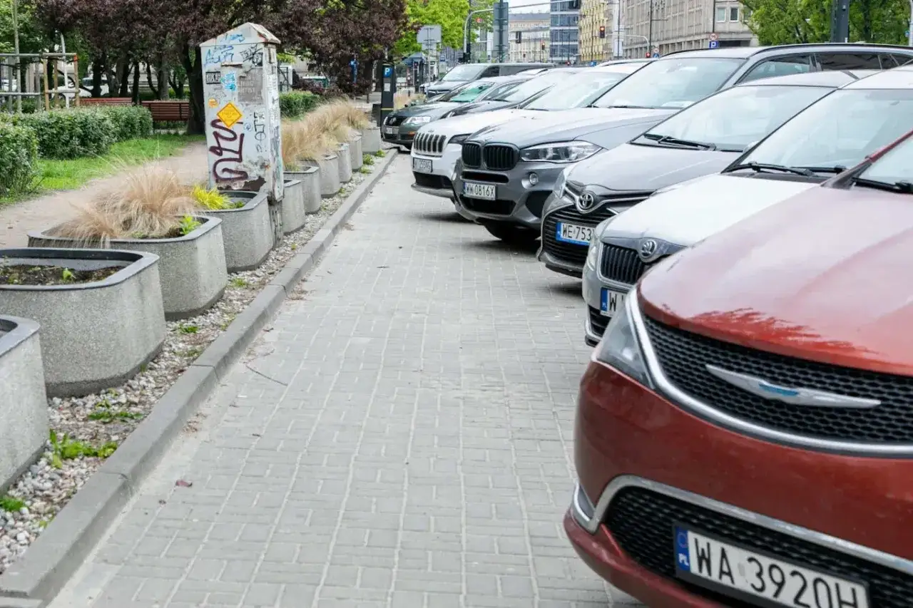 Samochody zaparkowane na chodniku, obok donic z trawą. Takie parkowanie na chodniku grozi mandatem.