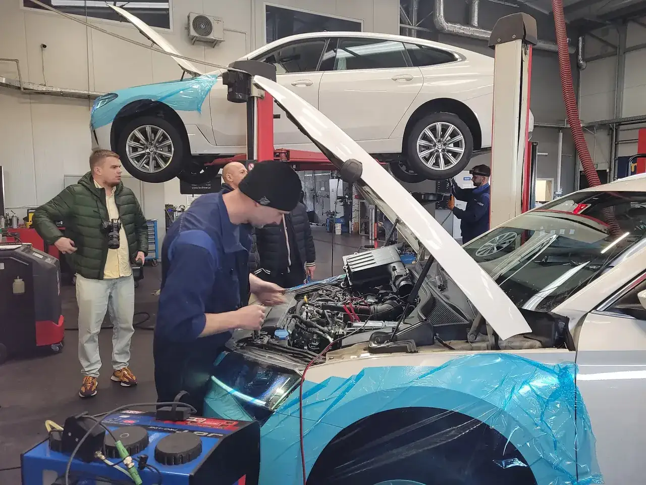 Mechanicy w warsztacie przeprowadzają płukanie silnika w samochodzie na podnośniku. Inny samochód również uniesiony.