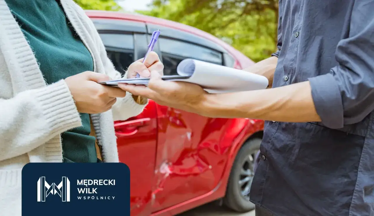 Osoba podpisuje dokumenty po wypadku samochodowym. Czerwony samoch&oacute;d ma widoczne uszkodzenia.