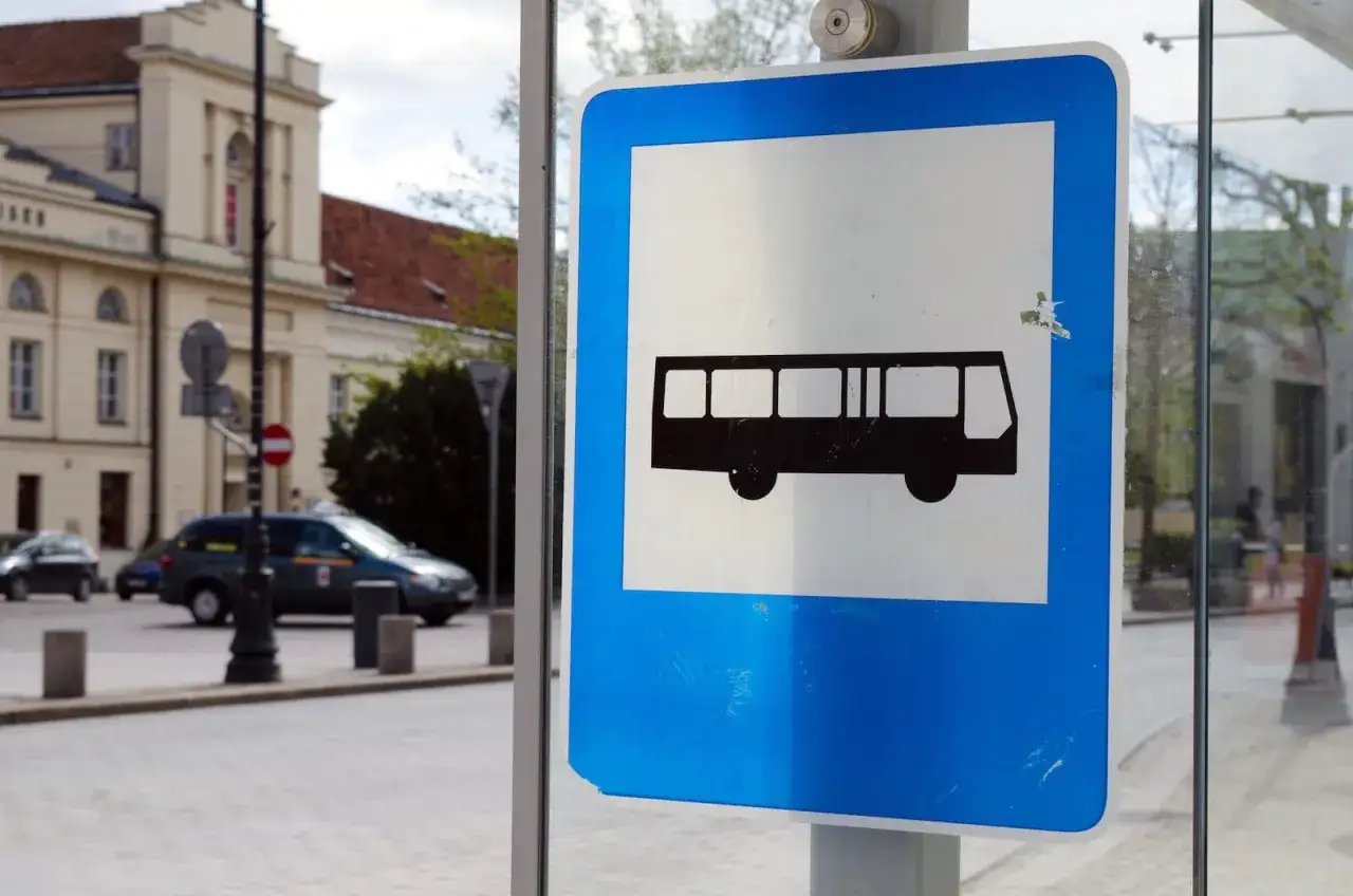 Znak przystanku autobusowego. Czy autobus ma pierwszeństwo? Na ulicy widać samochody i budynek.
