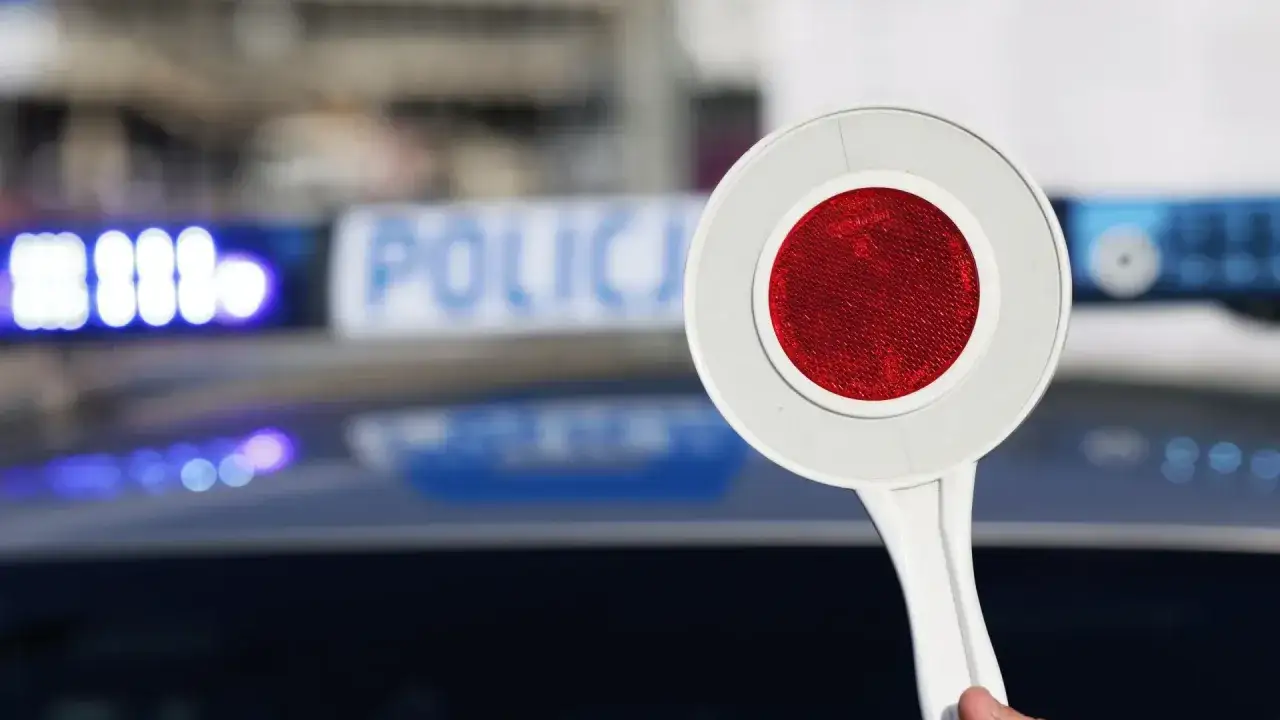 Tarcza policyjna z czerwonym odblaskiem, w tle niebieskie światła i napis 