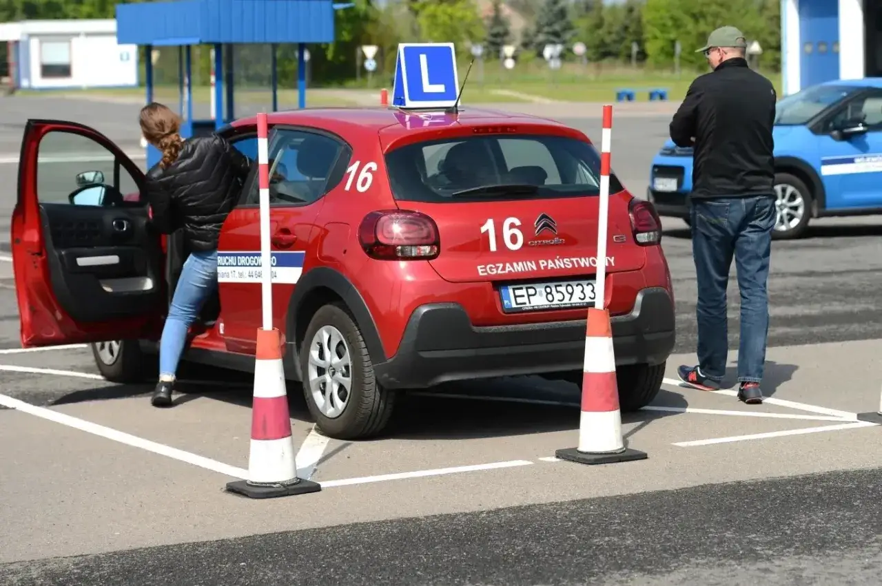 Na placu manewrowym młoda kobieta wsiada do czerwonego auta z literą