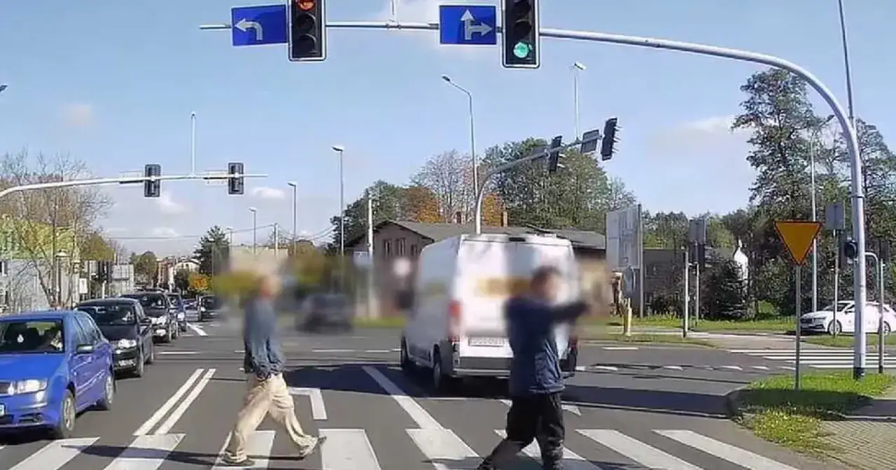 Zielone światła sygnalizacyjne pozwalają pieszym przejść przez pasy. Samochody stoją w korku.
