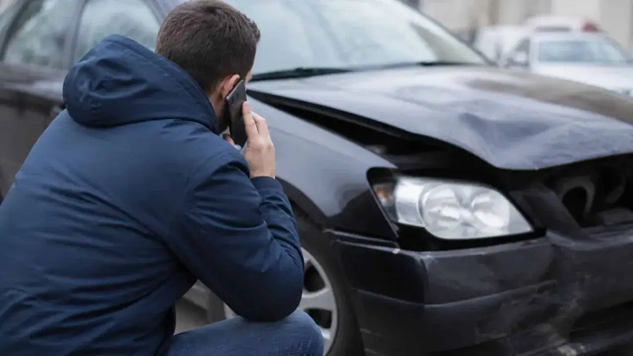 Mężczyzna rozmawia przez telefon obok uszkodzonego samochodu, martwiąc się o utratę wartości handlowej pojazdu po wypadku.
