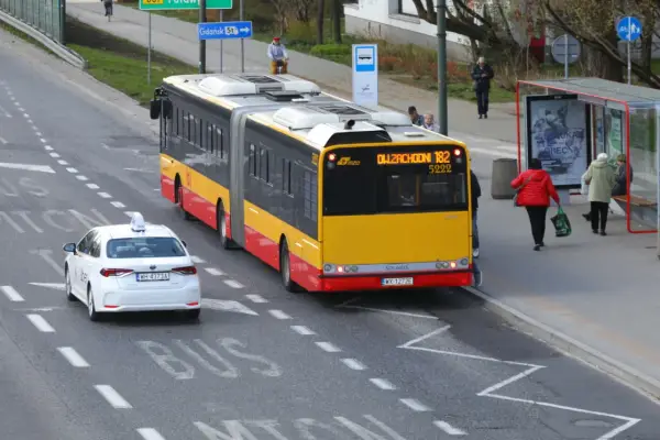 Żółto-czerwony autobus przegubowy linii 182 zbliża się do przystanku. Czy autobus ma pierwszeństwo przed białym autem taxi?
