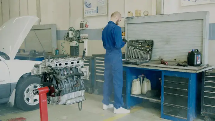 Mechanik w niebieskim kombinezonie przy silniku samochodu. Obok skrzynka z narzędziami i płyny. Lepiej zrobić przegląd, by uniknąć mandatu za brak przeglądu.