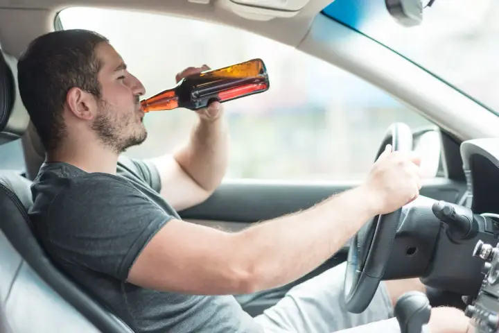 Mężczyzna pije piwo za kierownicą. Powyżej 0,5 promila jaka kara grozi za jazdę pod wpływem alkoholu.